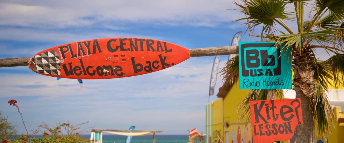 A beachside sign with 'PLAYA CENTRAL Welcome back' and 'Kite Lesson' signs.