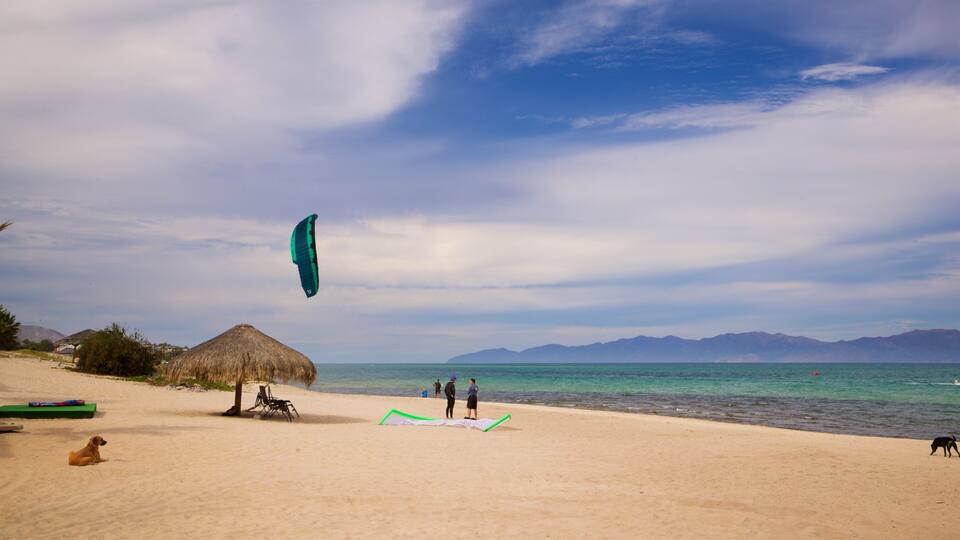 A beach with a thatched hut, people flying a kite, and mountains in the background.