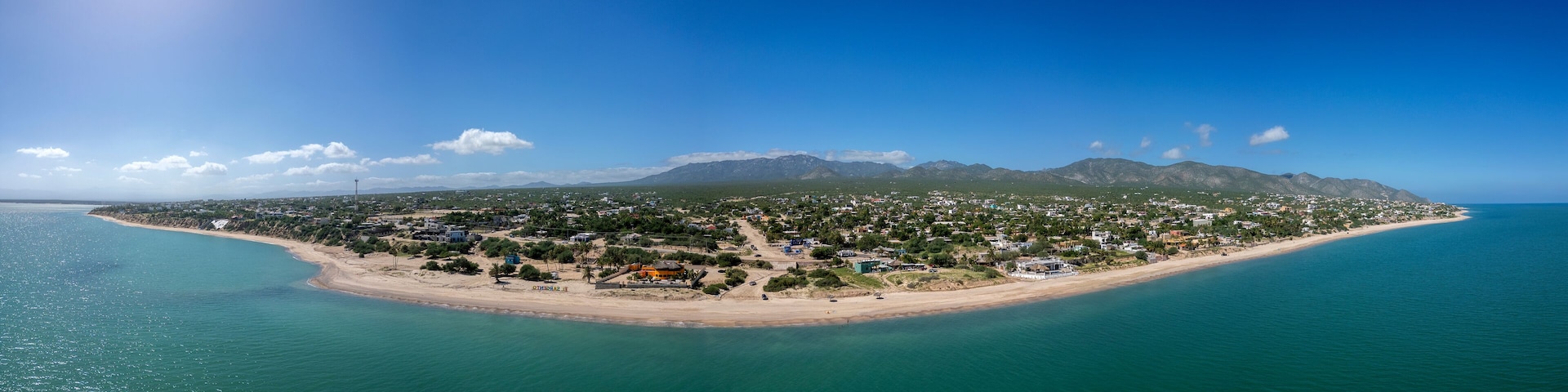 Vista aérea de un pueblo costero con playa, casas y montañas al fondo.