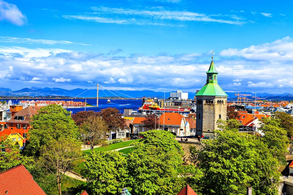 A coastal town with a prominent church tower, a bridge, and mountains in the background.
