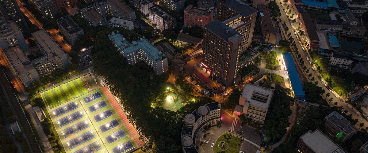 An aerial view of a cityscape at night with illuminated buildings, a sports field, and a curved structure with domes.