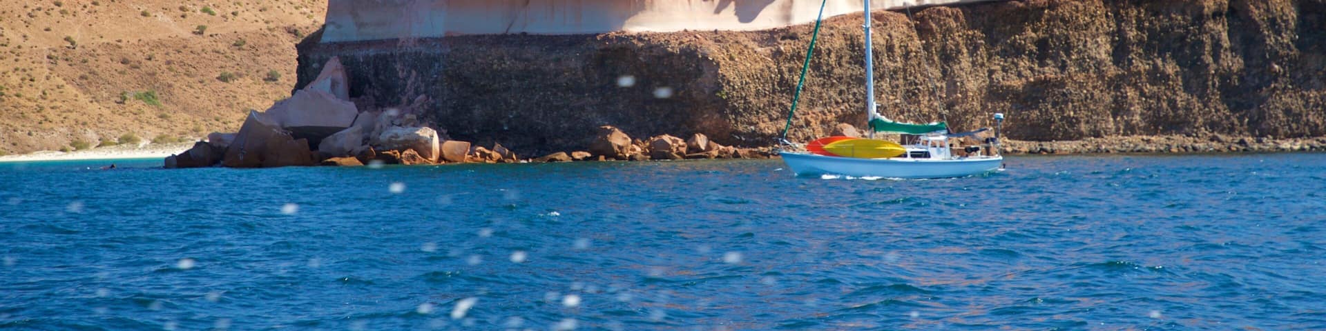 A rocky cliff by a clear blue sea with a sailboat anchored nearby.
