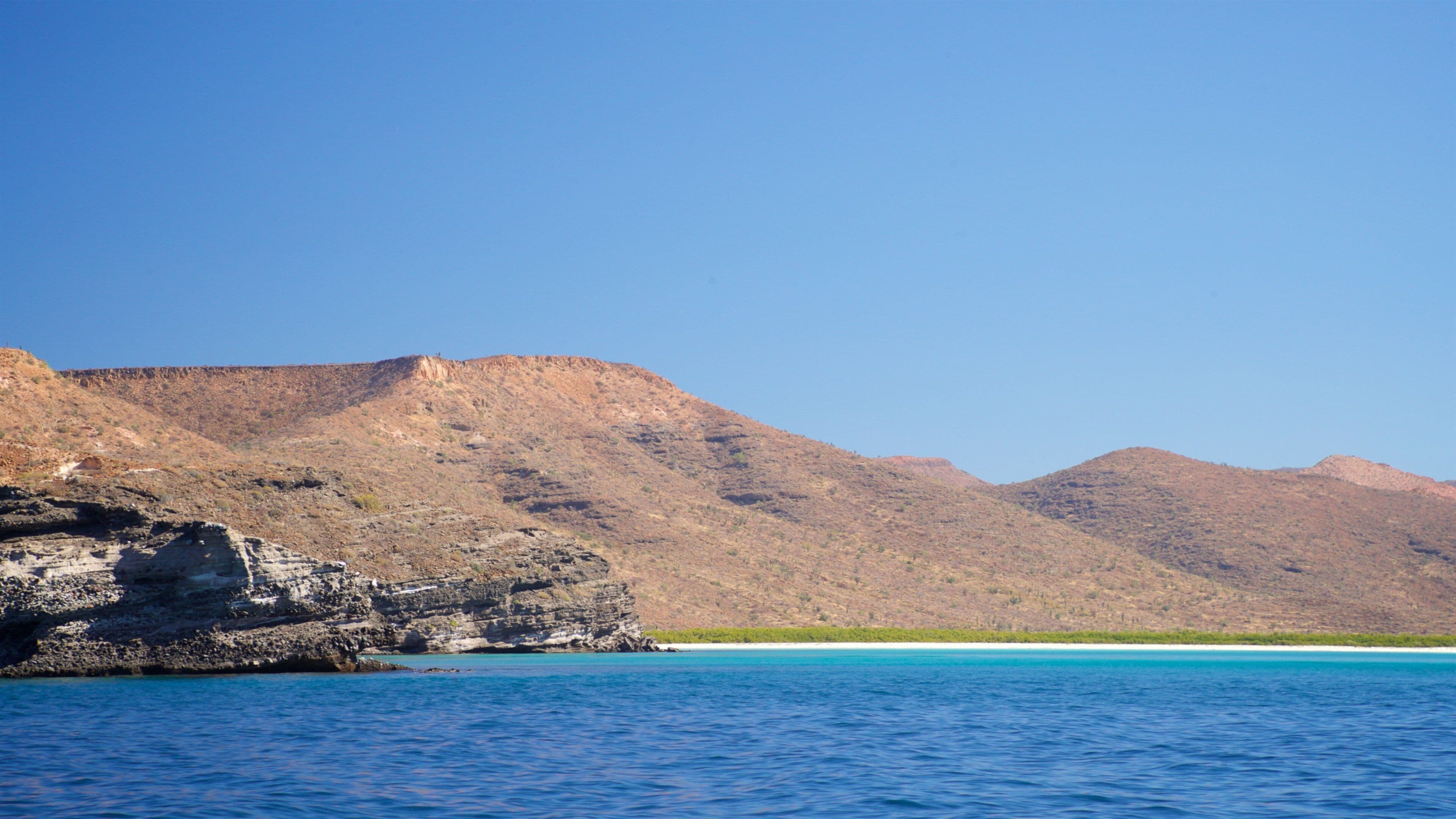 A coastal landscape with clear blue waters, rocky cliffs, and distant mountains under a clear sky.