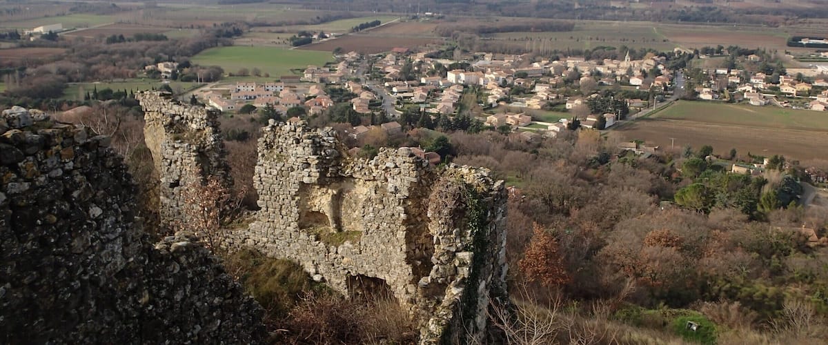 A historic stone castle ruins overlooking a rural village with fields and distant mountains.