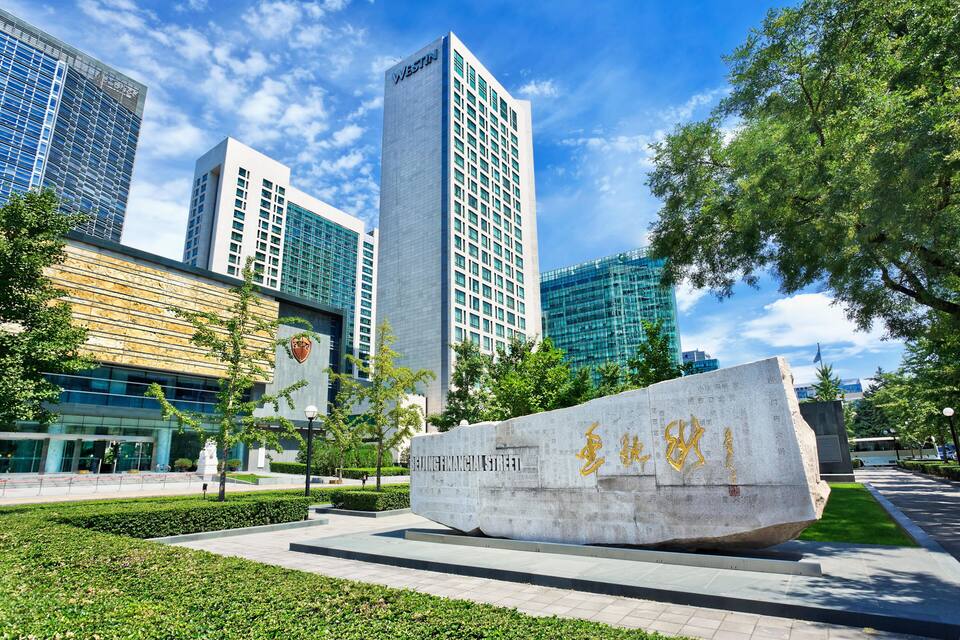 A modern urban plaza with a large stone monument, tall buildings, and green trees.