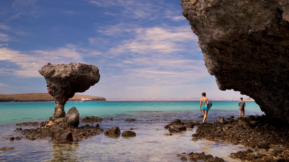 A rocky shoreline with clear turquoise water, a boat in the distance, and two people standing near a rock formation.