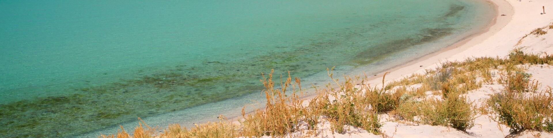 Un paisaje costero con una playa arenosa, aguas turquesas cristalinas y una colina rocosa al fondo.