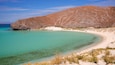 Un paysage côtier avec une plage de sable, une eau turquoise et limpide, et une colline rocheuse en arrière-plan.