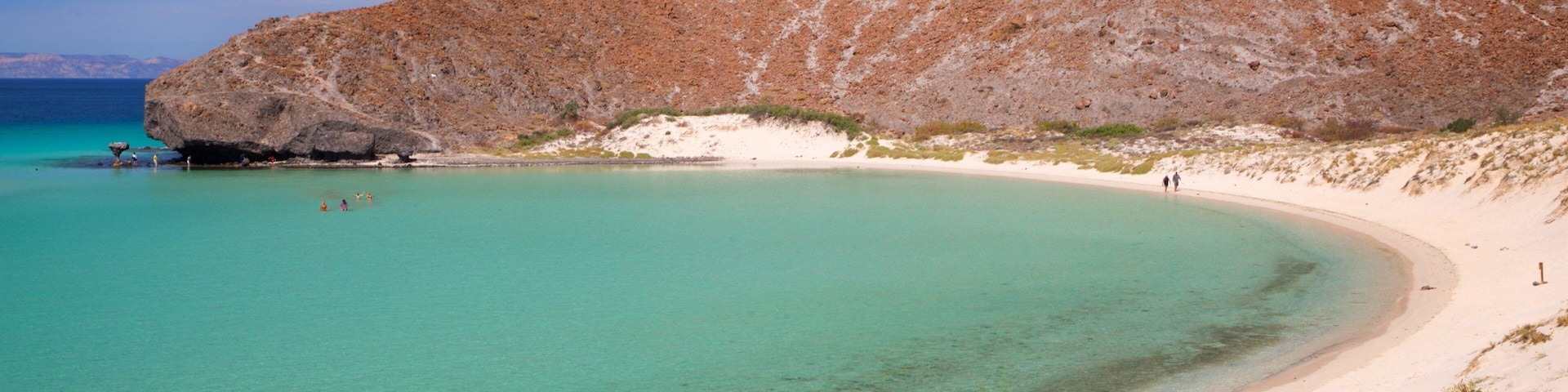 Paisagem costeira com uma praia arenosa, águas turquesa cristalinas e uma colina rochosa ao fundo.