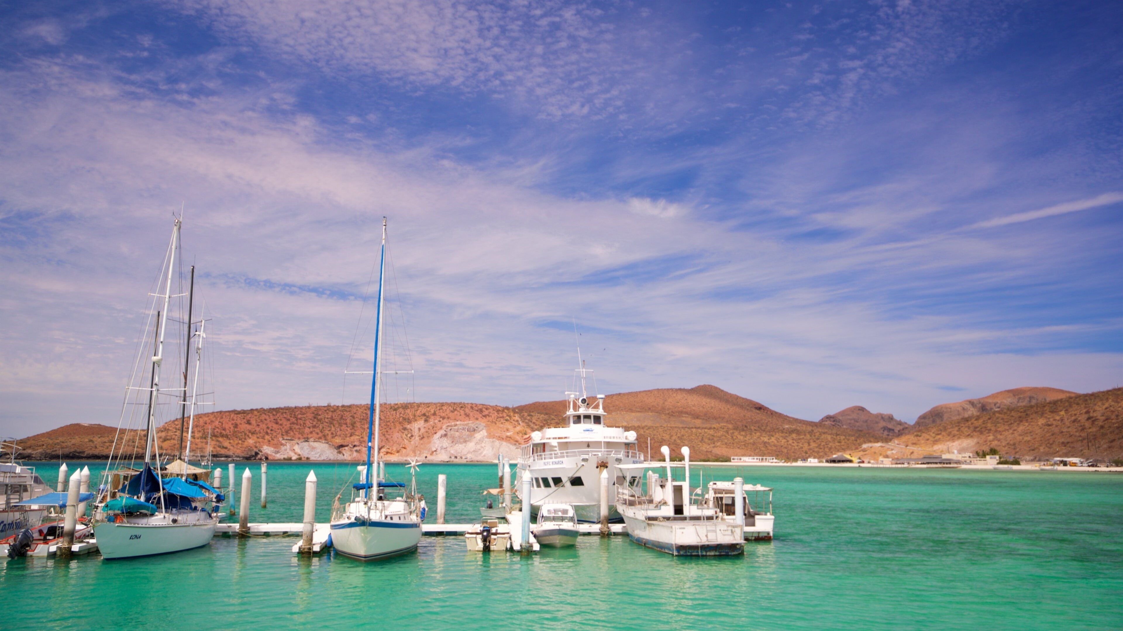A marina with several boats docked, surrounded by a clear turquoise sea and distant hills under a partly cloudy sky.