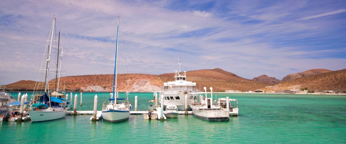 A marina with several boats docked, surrounded by a clear turquoise sea and distant hills under a partly cloudy sky.