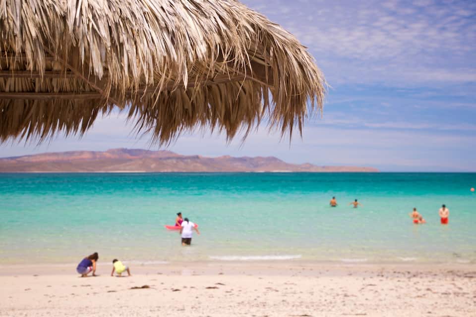 En strand med en byggnad som har ett tak av halmtak, kristallklart turkost vatten och berg i fjärran.