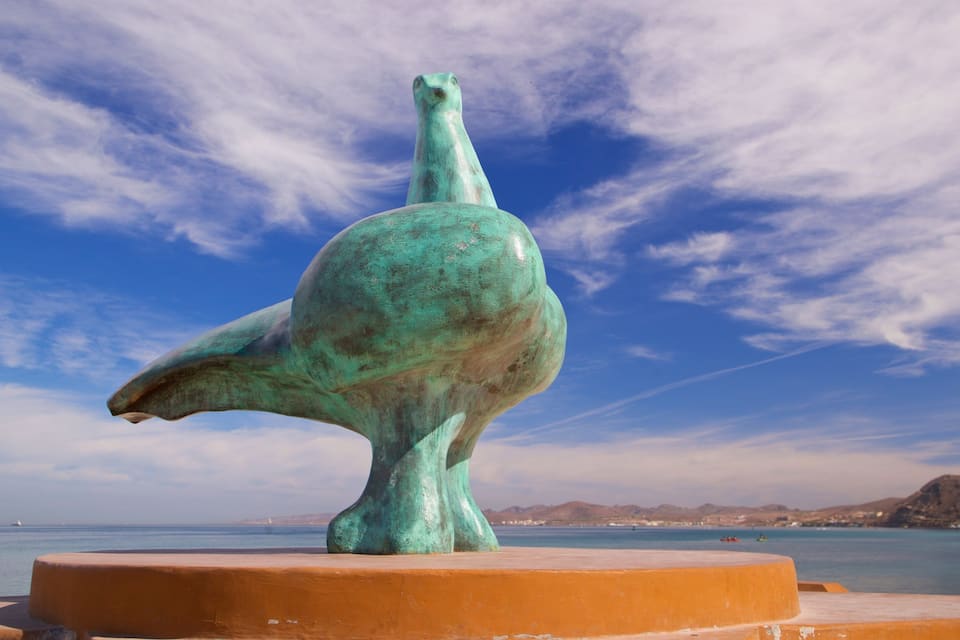 En bronzeskulptur af en fugl på en orange piedestal ved havet.