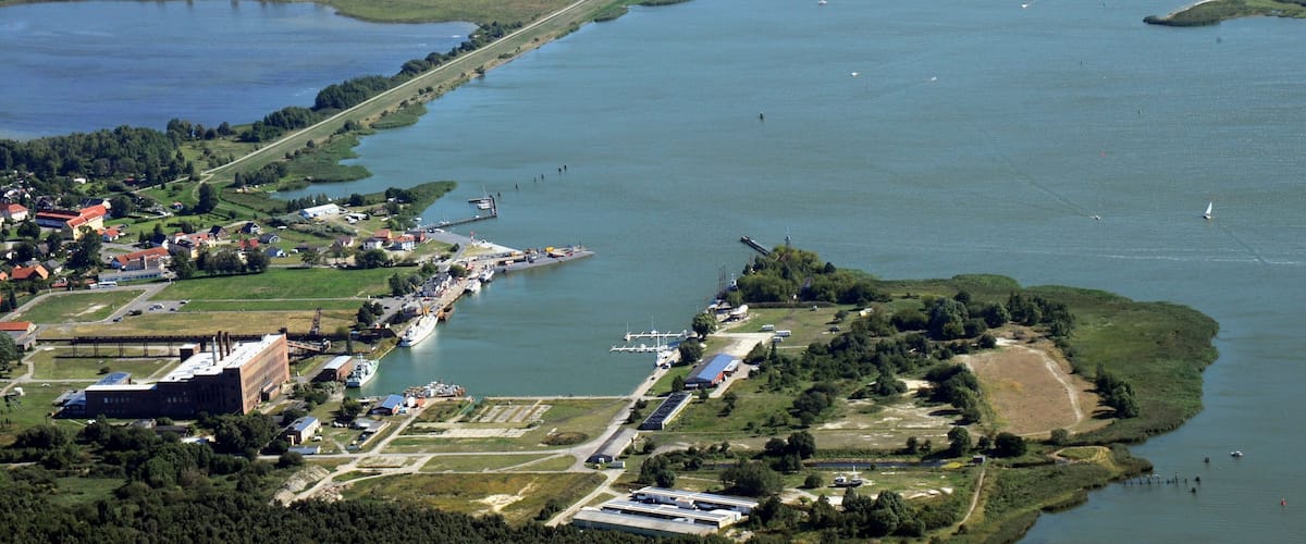 An aerial view of a coastal industrial area with a river, a marina, and a forested peninsula.