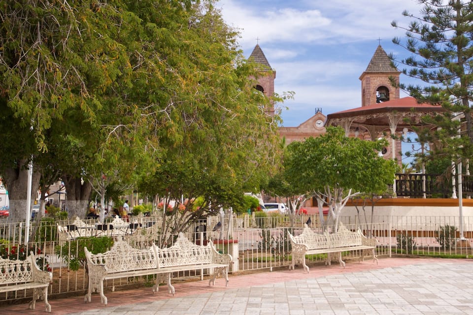 A plaza with ornate benches, a clock tower, and a church with two spires.