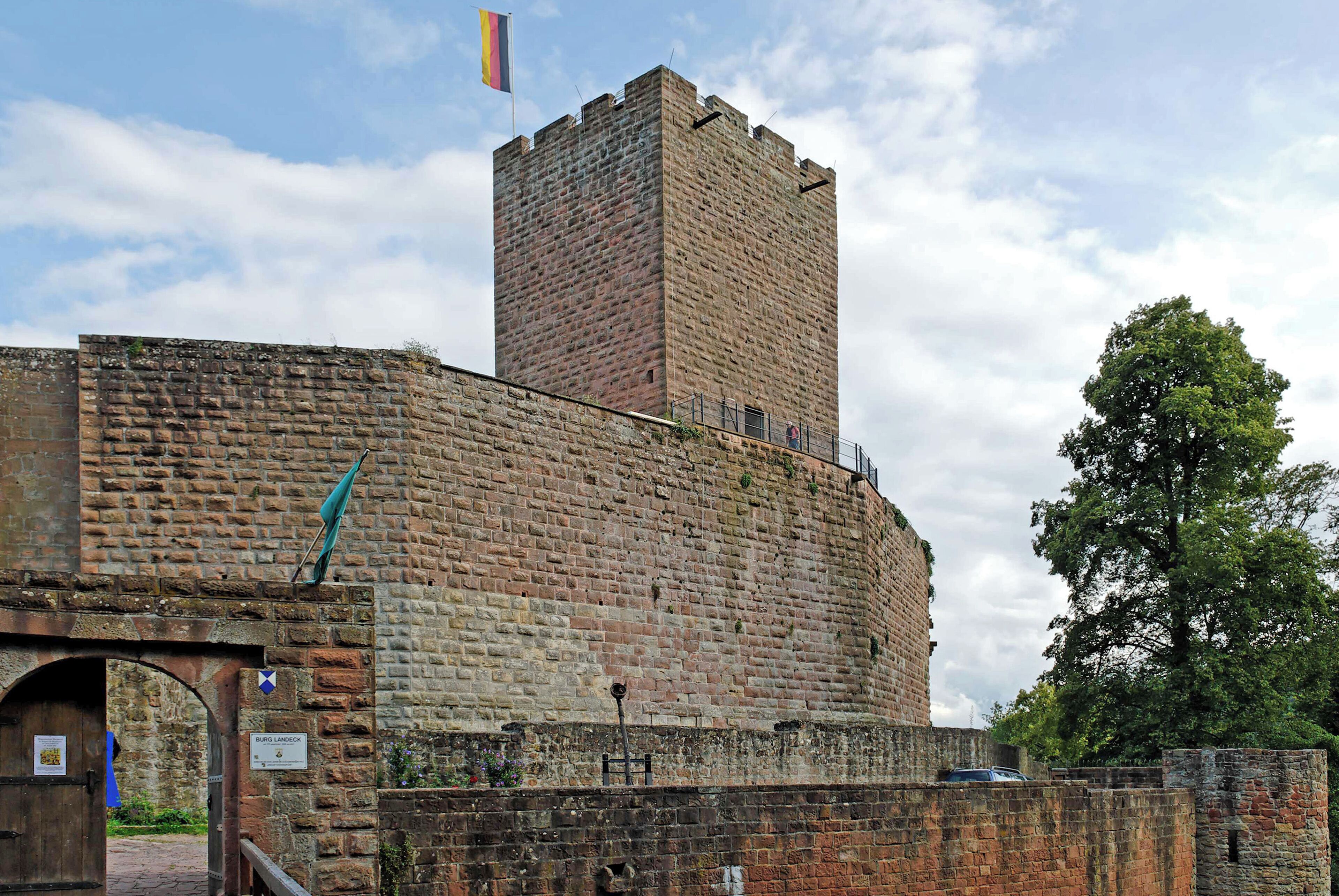 A historic brick tower with a flag, a gate, and a tree in the background.