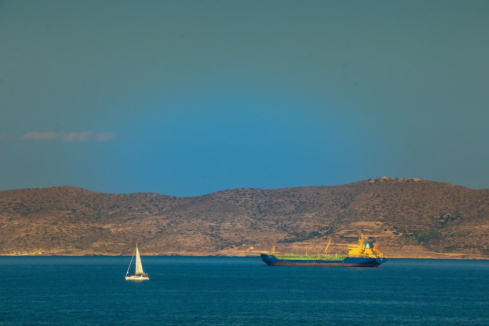 A sailboat and a cargo ship in the sea with a mountainous coastline in the background.