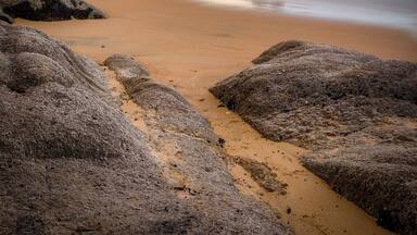 Uma praia com pedras grandes em primeiro plano e mar calmo.