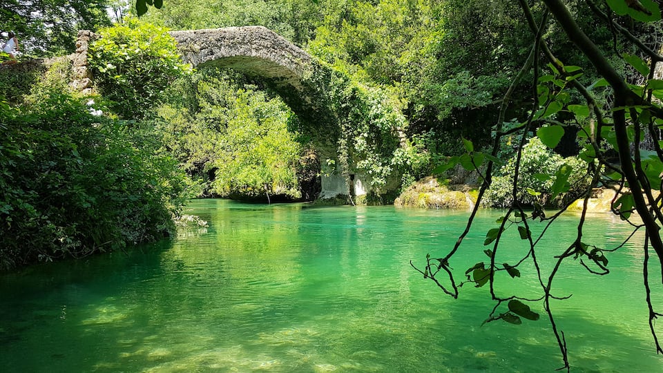 A river with clear water, surrounded by lush greenery and a stone bridge.