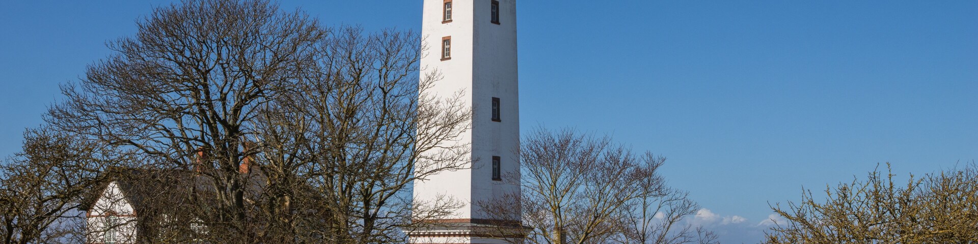 A white lighthouse with a red top, surrounded by trees and a hedge.
