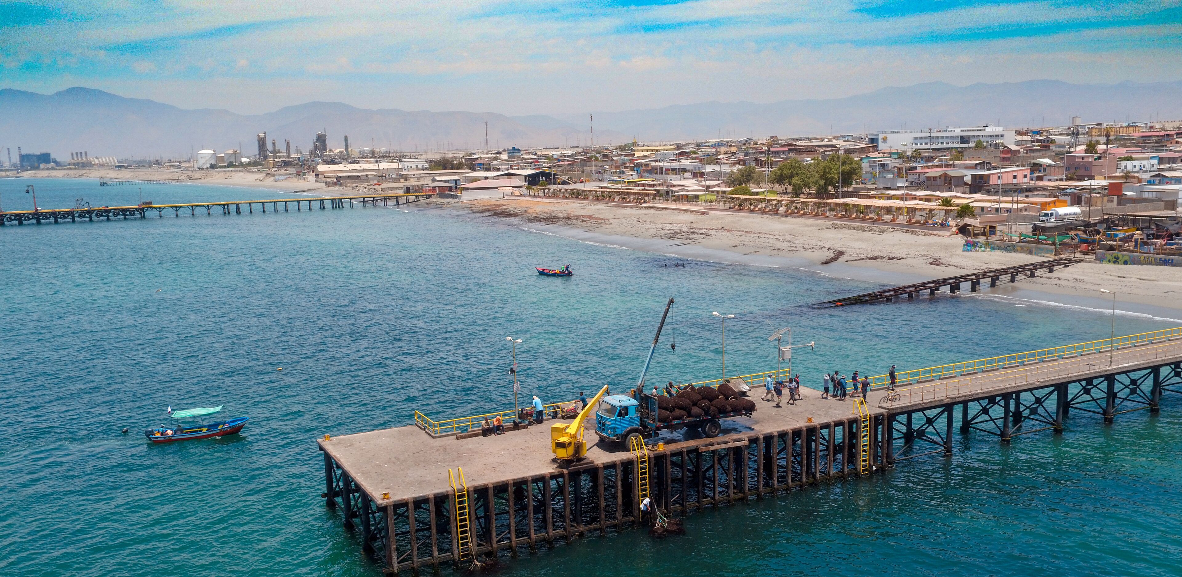 A pier extending into a calm sea with a small boat, a docked truck, and a distant cityscape.