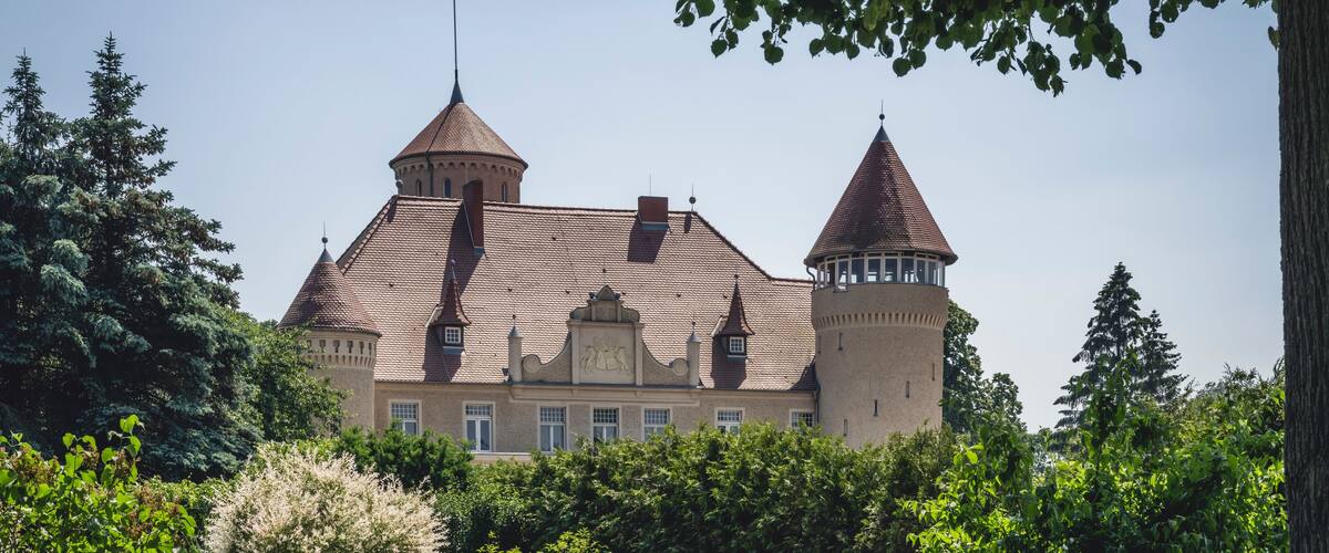 A castle with two towers and a central building surrounded by lush greenery.