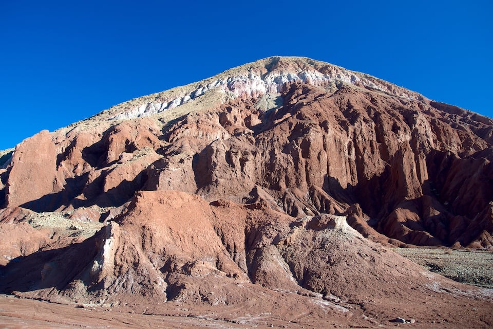 Montaña escarpada con formaciones rocosas de color marrón rojizo y vegetación escasa bajo un cielo azul despejado.