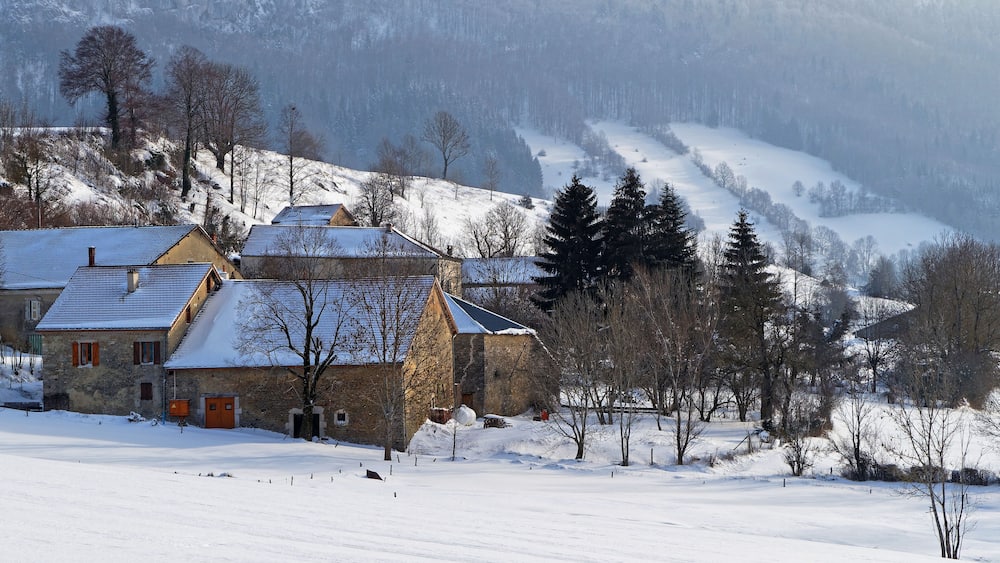 Un village enneigé, avec des maisons en pierre et une montagne boisée en arrière-plan.