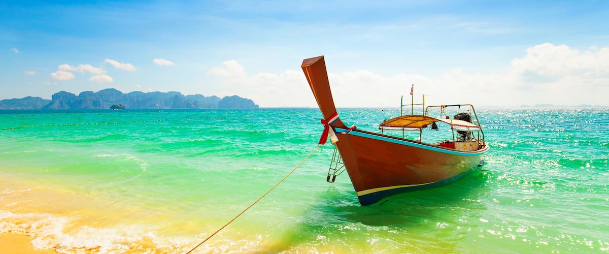 A wooden boat anchored near a beach with clear turquoise water and distant rocky islands.