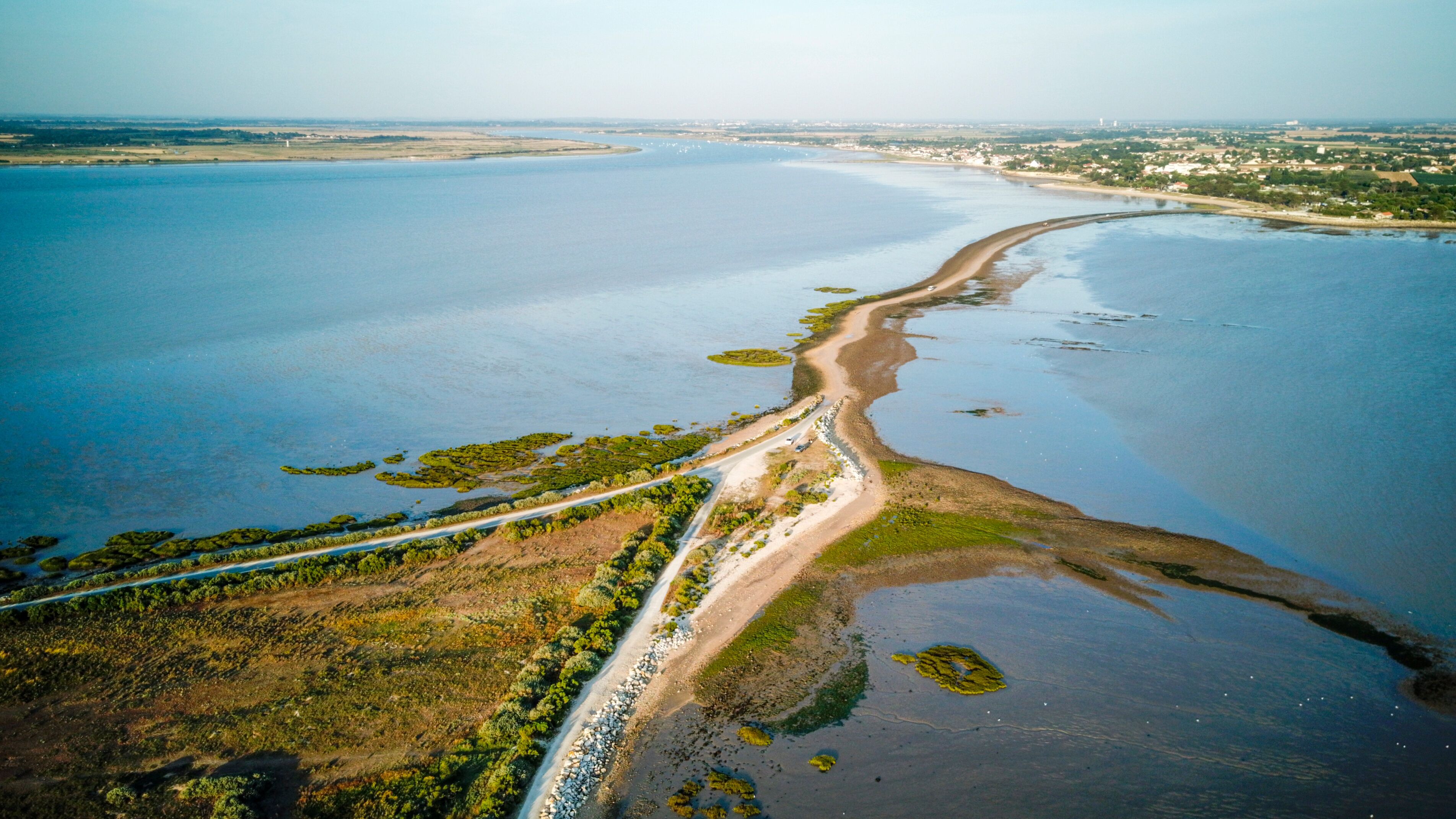 An aerial view of a coastal area with a sandy beach, a road running parallel to the shore, and patches of vegetation.