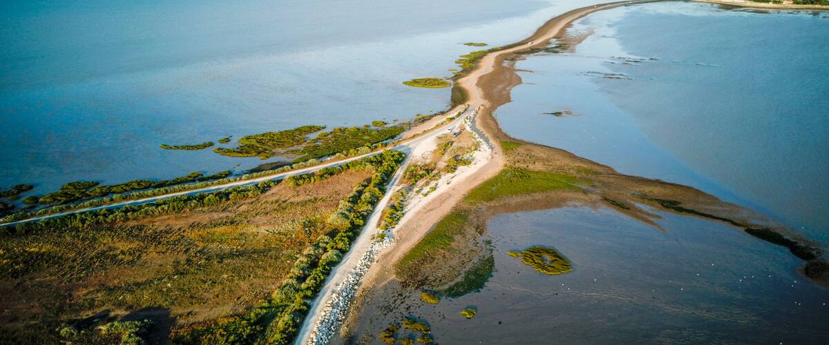 An aerial view of a coastal area with a sandy beach, a road running parallel to the shore, and patches of vegetation.