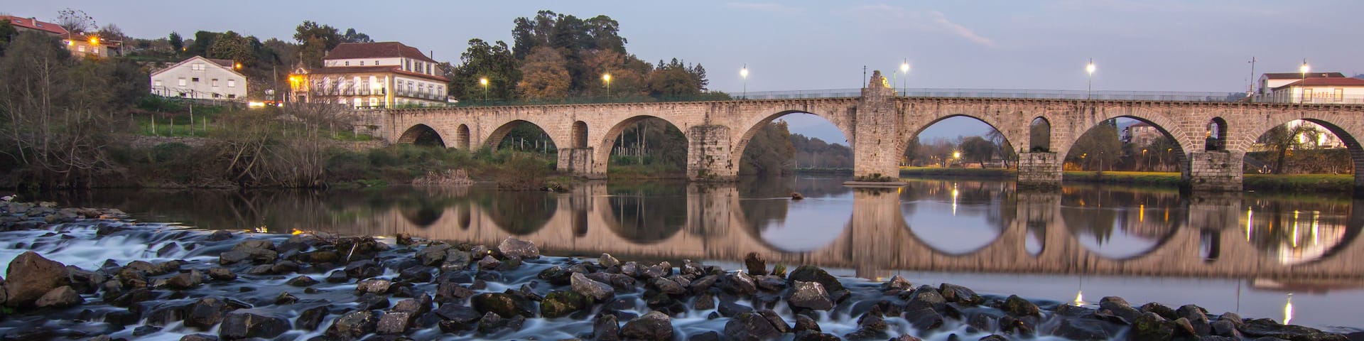 A stone bridge over a river with a reflection in the water, surrounded by trees and buildings.