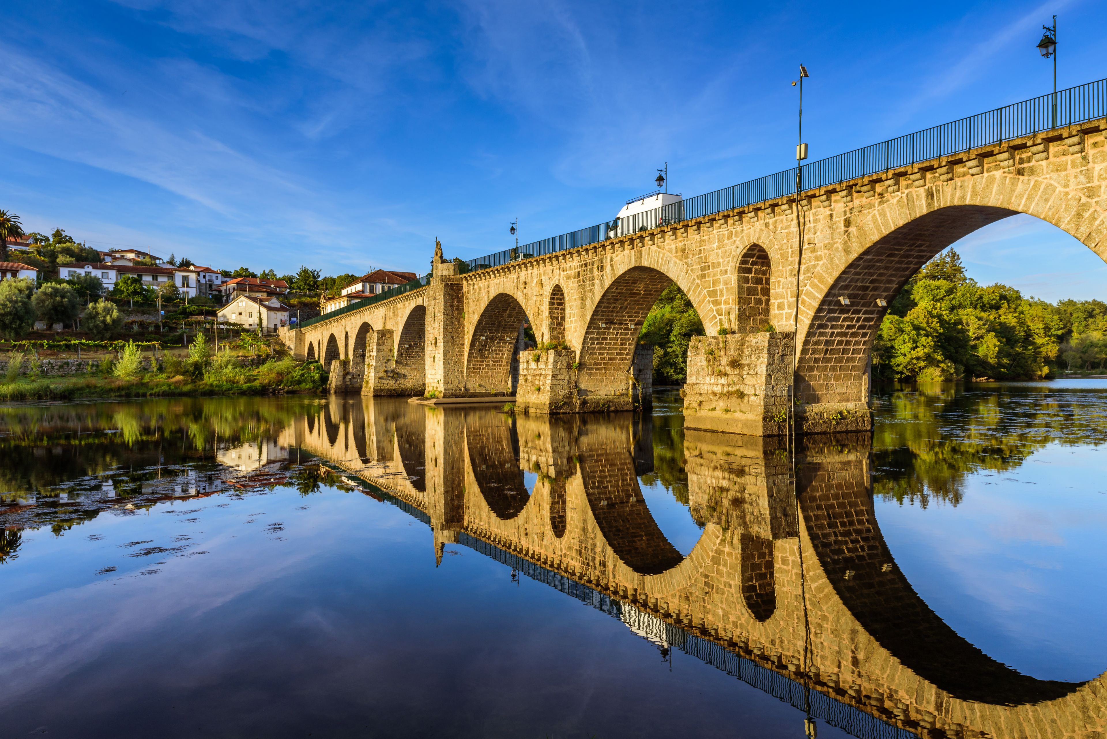 A stone bridge with multiple arches over a calm river, reflecting the structure and surrounding greenery.