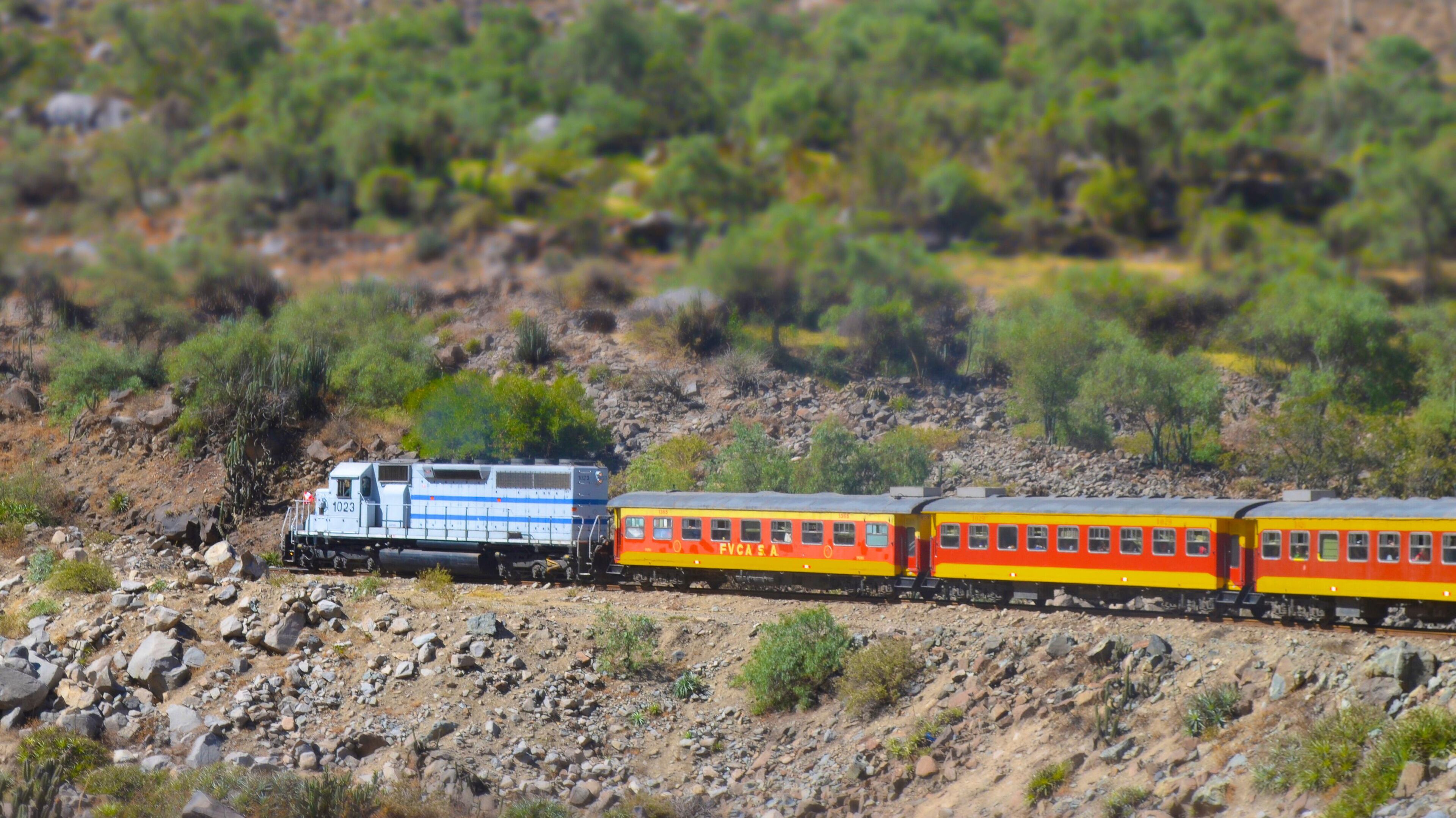 A train with a locomotive and passenger cars traveling through a rocky, hilly landscape with sparse vegetation.