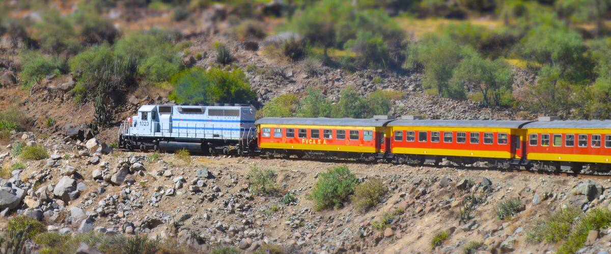 A train with a locomotive and passenger cars traveling through a rocky, hilly landscape with sparse vegetation.