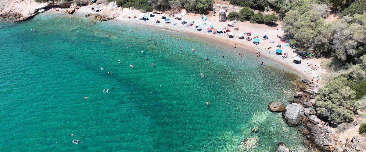 A coastal beach with clear turquoise waters, rocky outcrops, and a line of beach umbrellas.