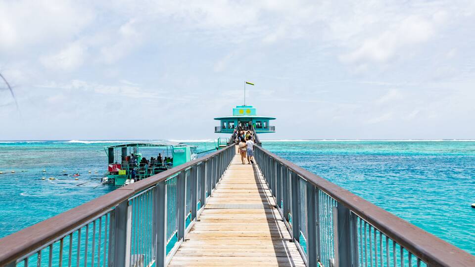 A wooden pier leading to a turquoise building with a flag, surrounded by clear blue water.
