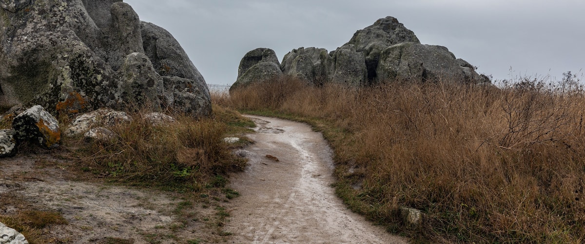 A dirt path flanked by large boulders and dry grass.