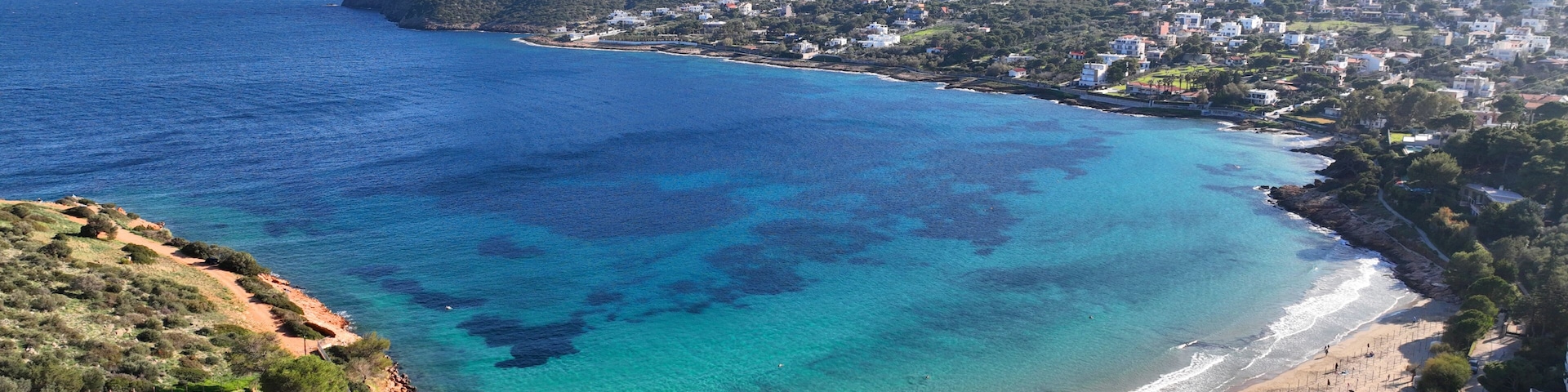 Uma cidade costeira com mar azul cristalino, uma praia de areia e montanhas ao fundo.
