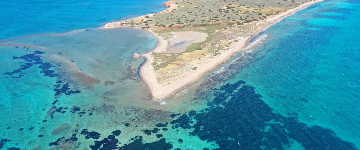 An aerial view of a coastal island with clear blue waters, sandy beaches, and scattered vegetation.