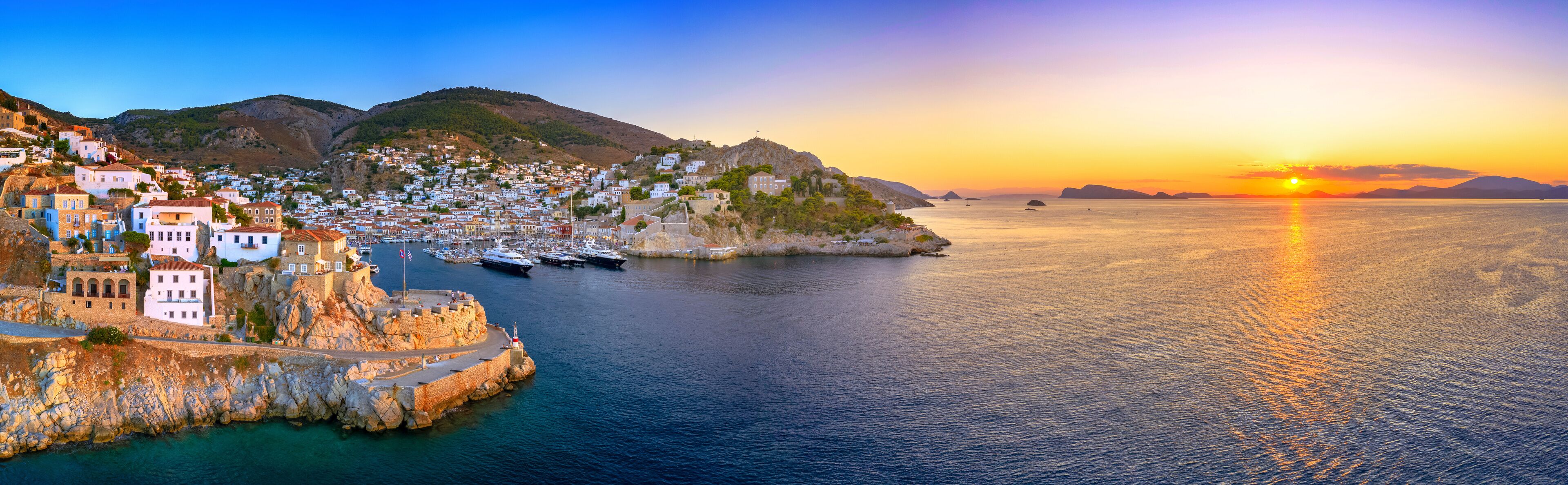 A coastal town with white buildings, a harbor with boats, and a sunset over the sea.