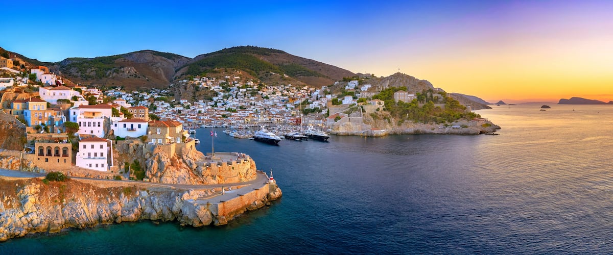 A coastal town with white buildings, a harbor with boats, and a sunset over the sea.