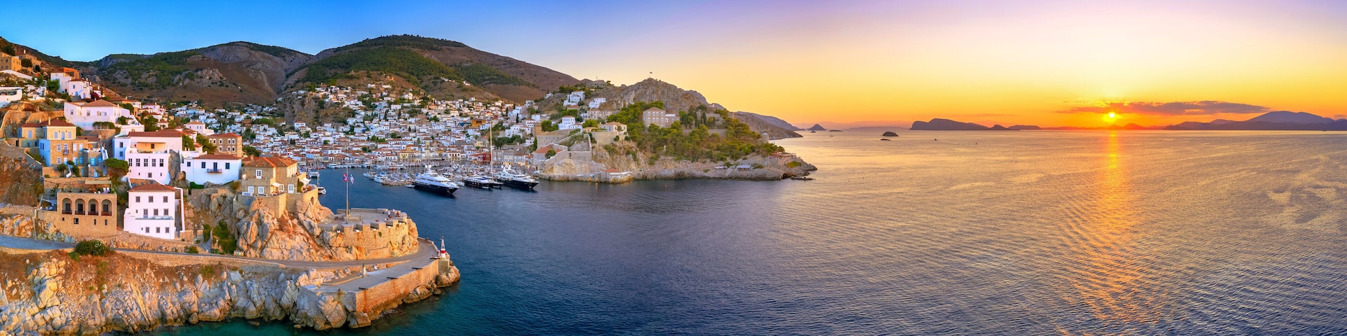 A coastal town with white buildings, a harbor with boats, and a sunset over the sea.