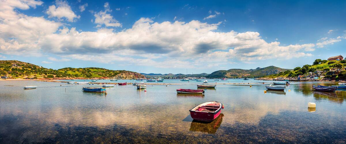 Uma baía calma com vários barcos ancorados, rodeada por colinas verdes e um céu azul e límpido.