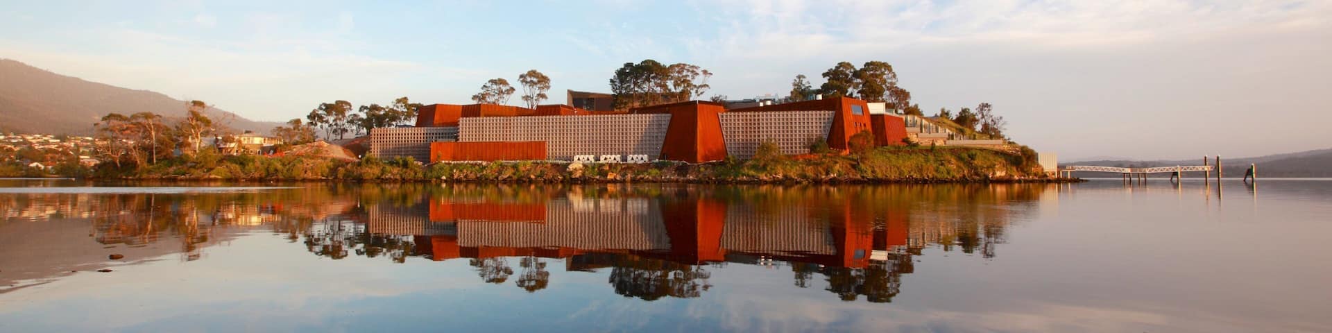 A modern building complex by a calm lake with reflections, surrounded by trees and hills.