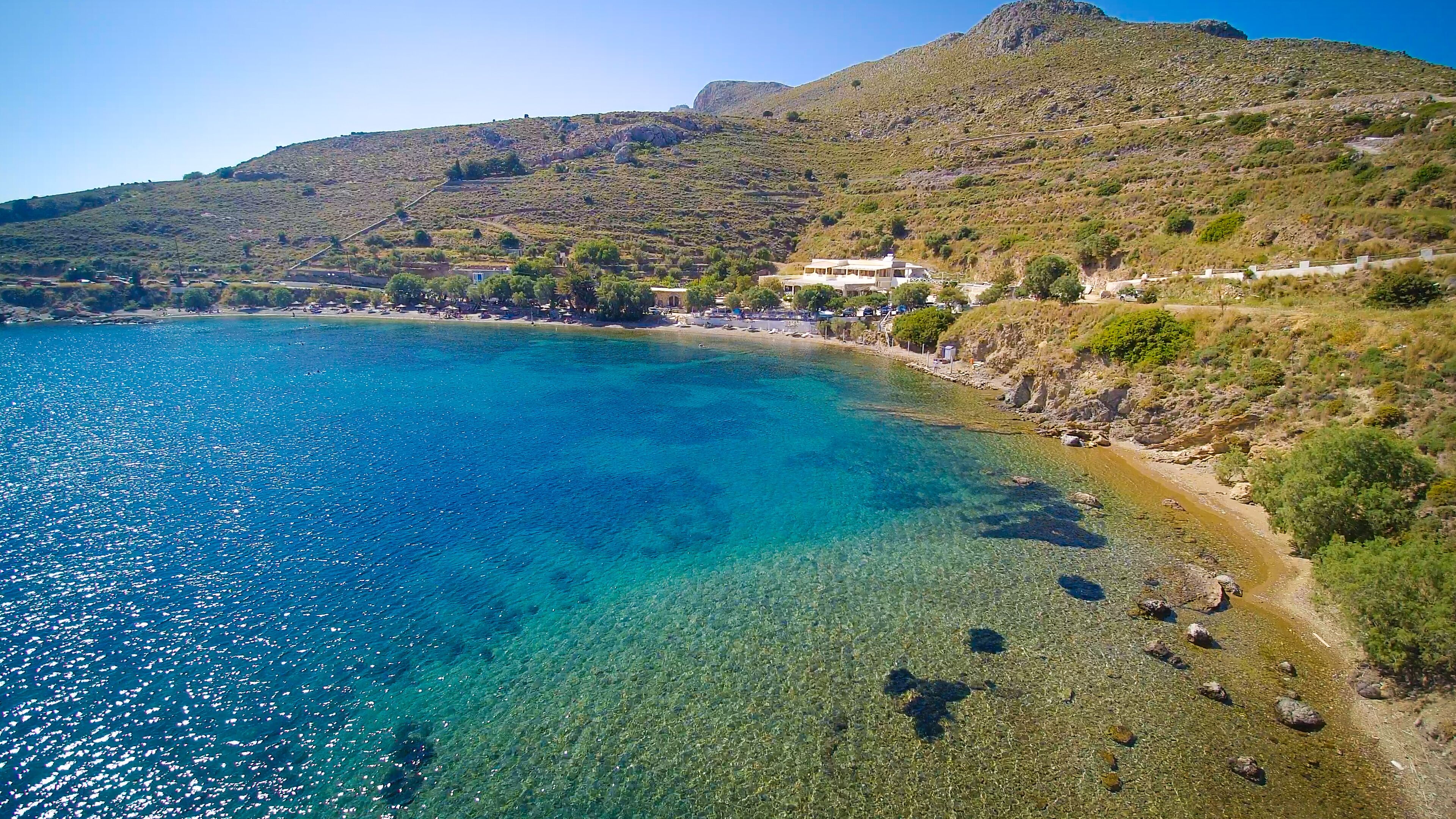 A coastal landscape with clear blue waters, rocky shores, and a hillside with sparse vegetation.