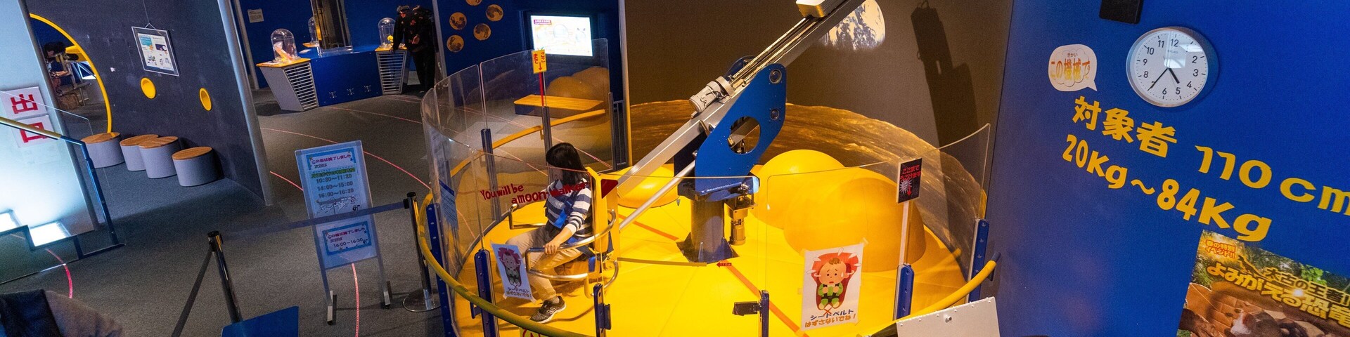 Exposição espacial com um grande modelo amarelo de estação espacial, uma criança manuseando um painel de controle e placas informativas em um ambiente azul.