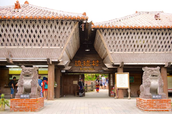 Entrada do Ryukyu Mura, com arquitetura tradicional, estátuas de leões de pedra e visitantes.
