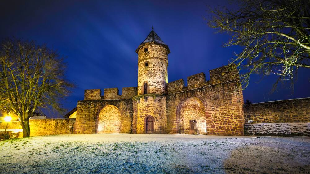 A historic castle with a tall tower, arched windows, and a snow-covered courtyard at night.