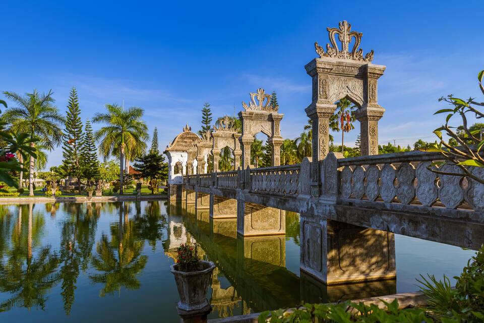 Eine Steinsche Brücke über ruhigem Wasser, mit verzierten Säulen und Palmen im Hintergrund.
